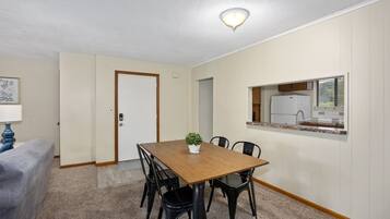 Un comedor con una mesa y sillas de madera, una planta pequeña en maceta y una cocina visible a través de una ventana.