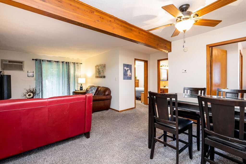 A room with a red sofa, wooden ceiling beams, a ceiling fan, a dining table with chairs, and a kitchen area visible through an open door.