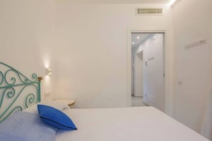A bedroom with a bed, a bedside table, a wall-mounted light, and a door leading to another room.