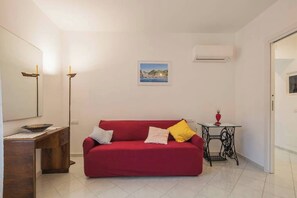 A red sofa with pillows, a wooden side table, a small table with a lamp, a framed picture on the wall, and an air conditioning unit.
