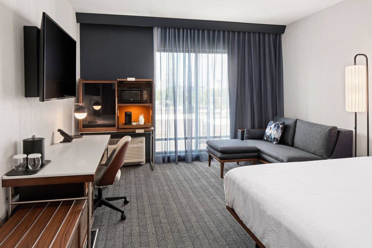Une chambre d’hôtel comprenant un lit, un bureau avec un ordinateur, un canapé, un téléviseur fixé au mur, un four à micro-ondes et une lampe.