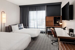 Une chambre d’hôtel avec deux lits, un bureau, une chaise, une télévision et une grande fenêtre avec des rideaux.
