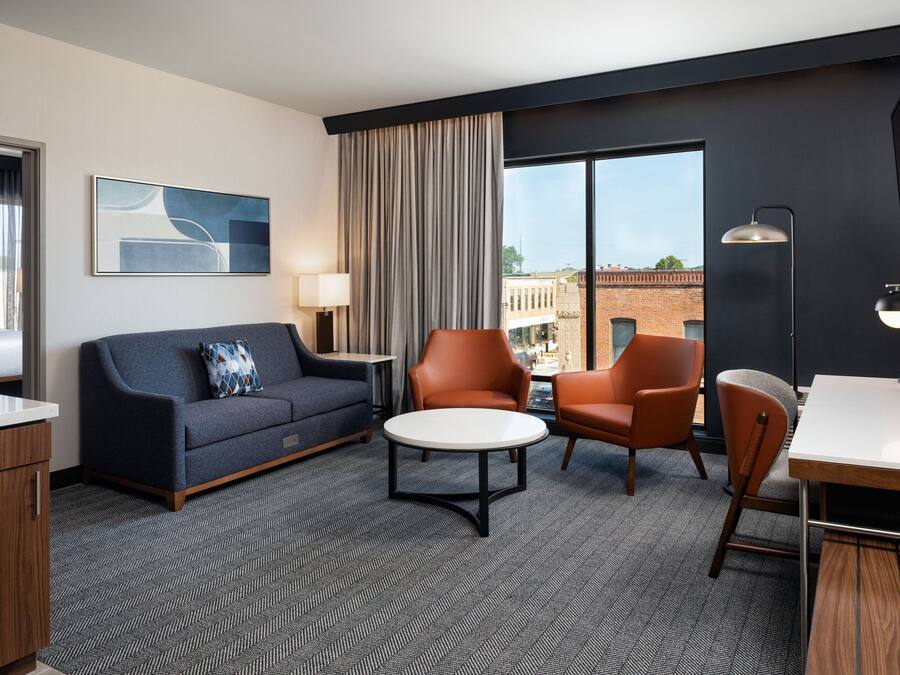 A modern hotel room with a sofa, two armchairs, a small round table, a desk, and a large window offering a city view.