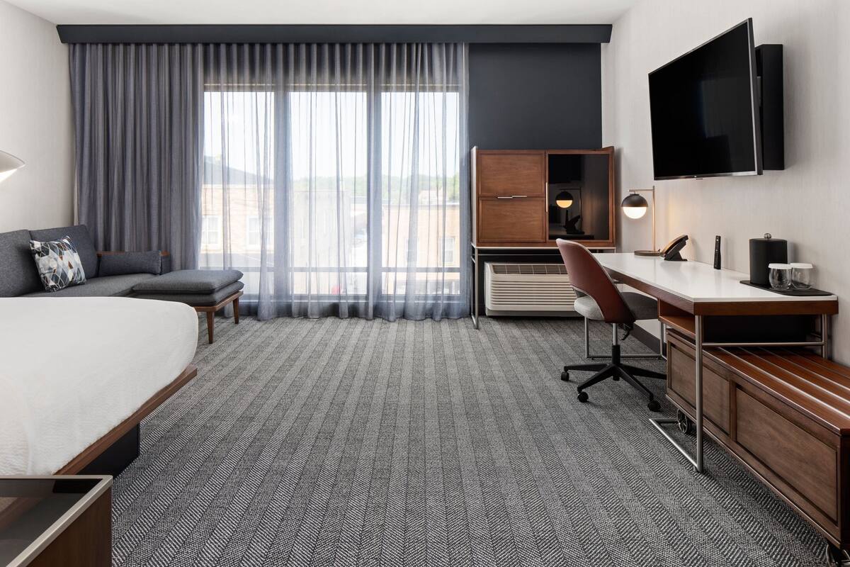 Une chambre d’hôtel avec un grand lit, un bureau, une chaise, une télévision et une fenêtre avec des rideaux.