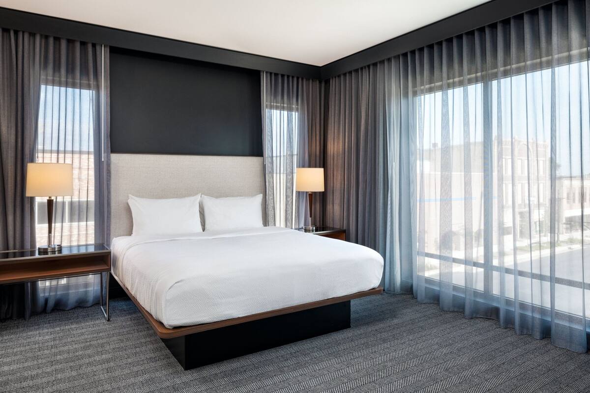 Une chambre d’hôtel moderne dotée d’un grand lit, de deux tables de chevet avec des lampes et de grandes fenêtres du sol au plafond, agrémentées de rideaux légers.