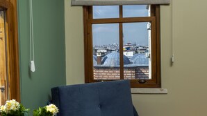 Ein Zimmer mit einem blauen Sofa, einem kleinen Tisch mit Blumen in einer Vase, einem Fenster mit Blick auf Gebäude und einem Vorhang.