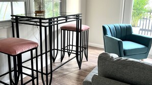A modern living room with a glass-top coffee table, two bar stools, a teal armchair, and a grey sofa.
