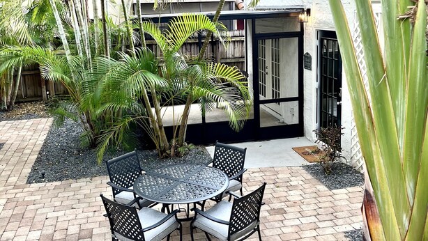 A patio area with a round table and chairs, surrounded by lush greenery and a brick floor.