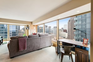 A modern living room with a grey sofa, a wooden desk, and a view of the cityscape through large windows.