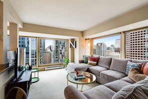 A modern living room with a sectional sofa, a glass coffee table, and a flat-screen TV. Large windows offer a city view.