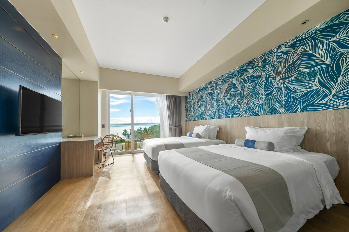 A modern hotel room with two beds, a TV, a balcony with a view, and a tropical leaf-patterned wall.