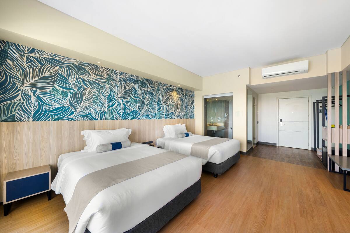 A hotel room with two beds, a large leaf-patterned wall, a wooden floor, a white door, and a small table with a mirror.