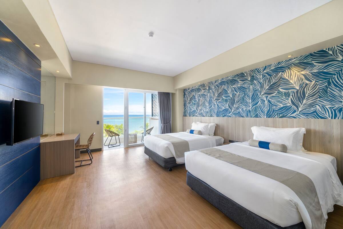 A modern hotel room with two beds, a desk, and a large window offering a view of the ocean.