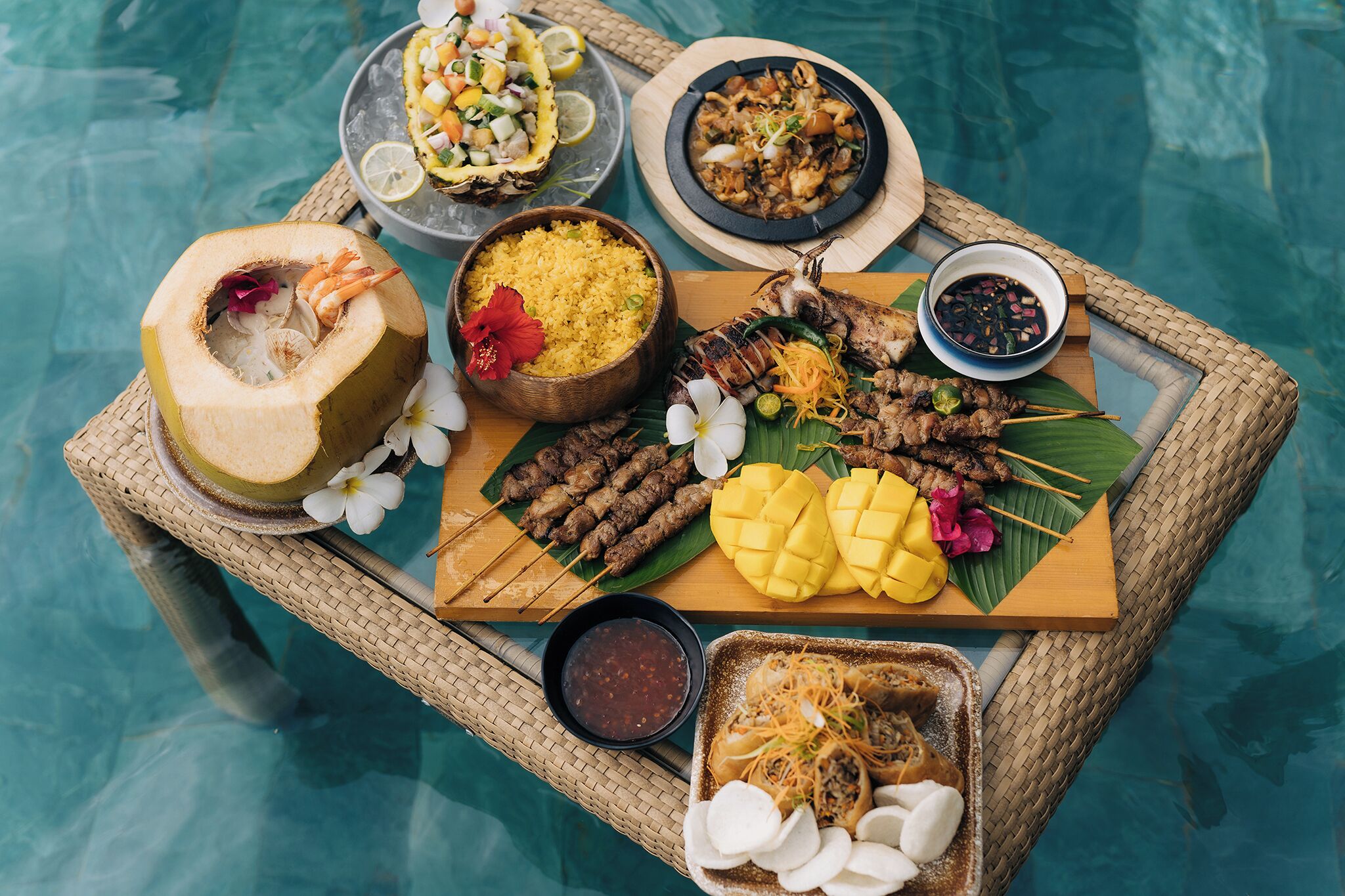 A variety of dishes including grilled meats, mango, and coconut drinks, served on a floating tray over a pool.