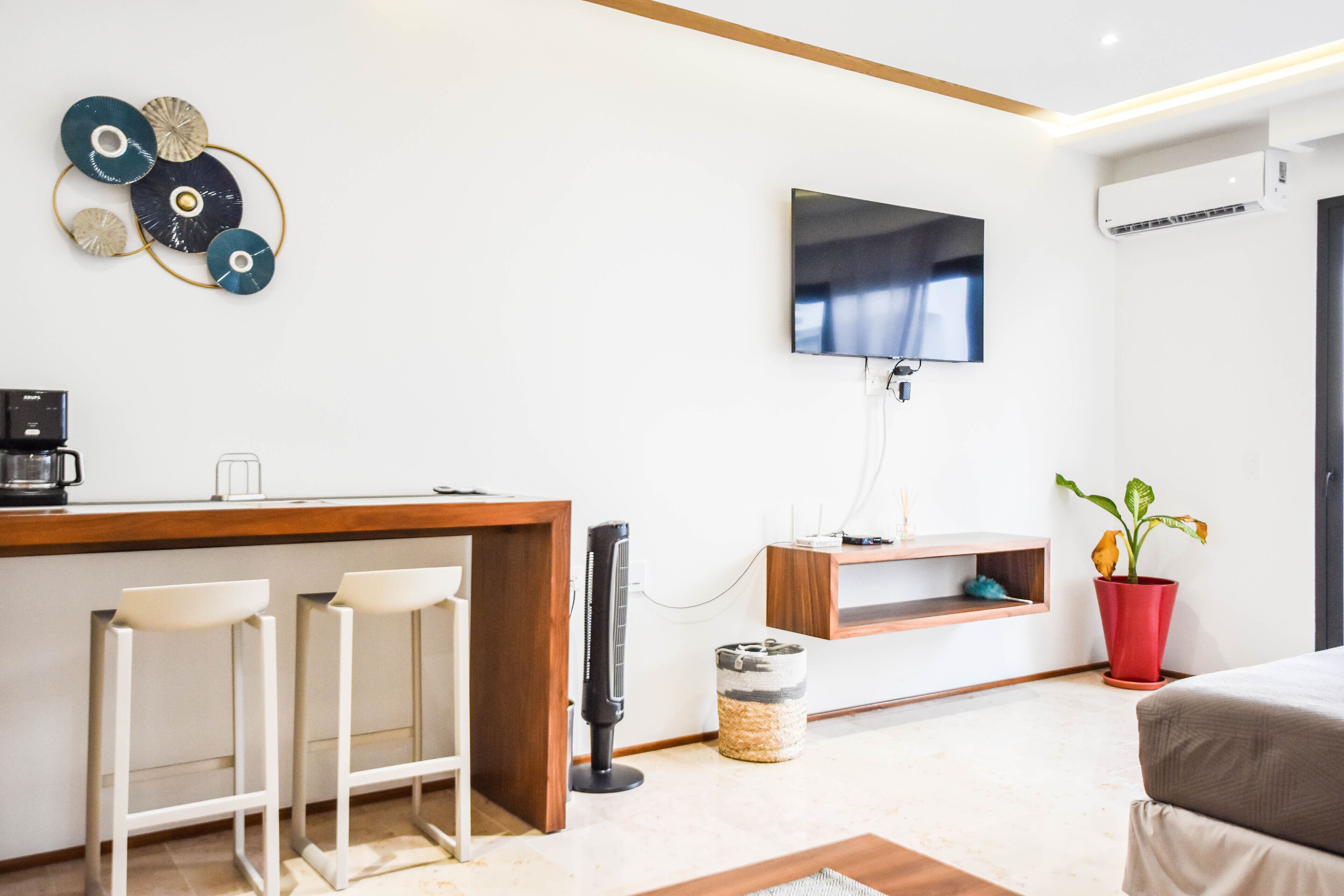 Een moderne woonkamer met een houten salontafel, twee witte barkrukken, een flatscreen televisie aan de wand, een rode potplant en een klein plankje met een decoratief object.