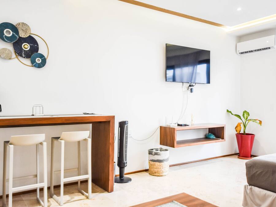 Een moderne woonkamer met een houten salontafel, twee witte barkrukken, een flatscreen televisie aan de wand, een rode potplant en een klein plankje met een decoratief object.
