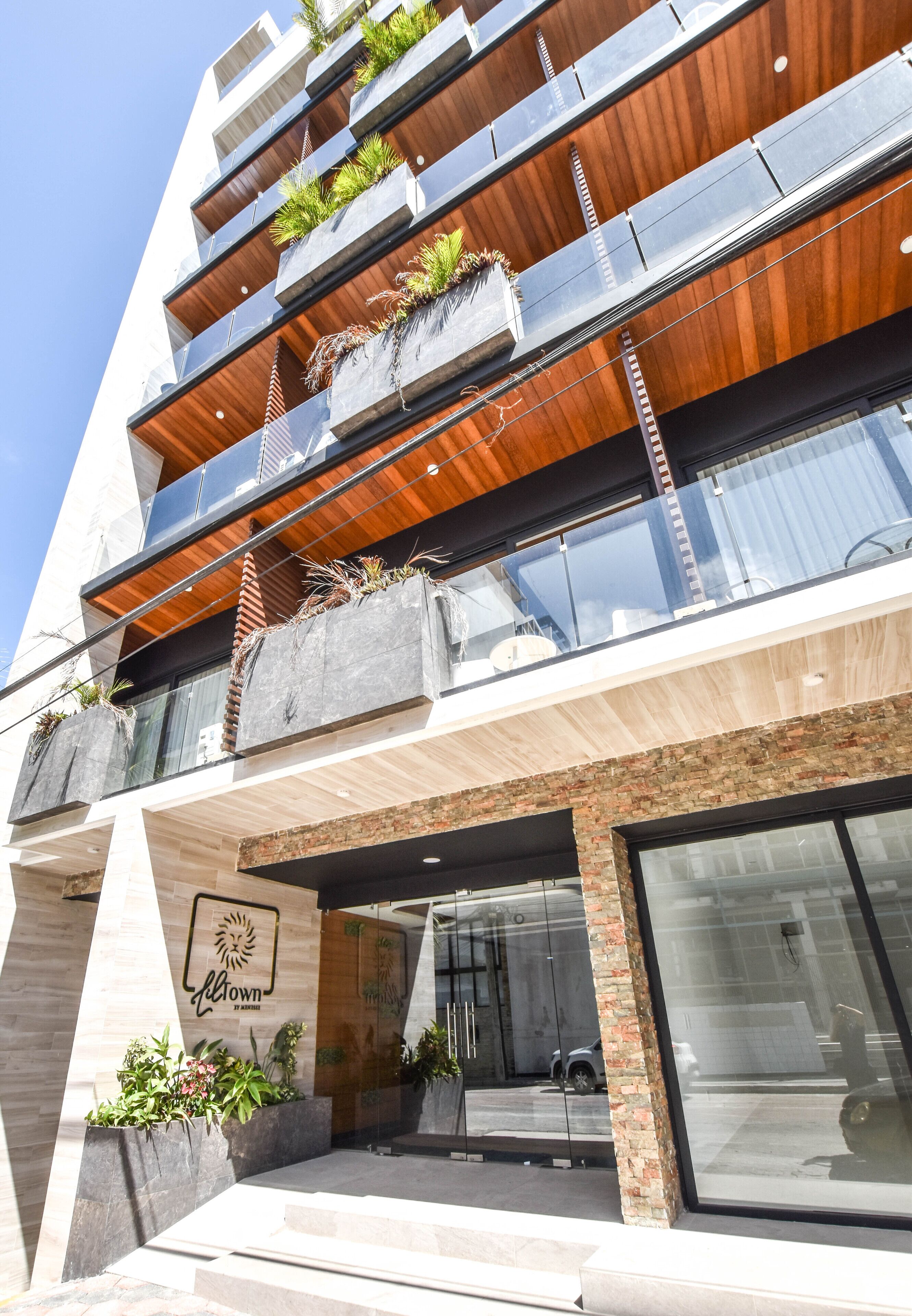 A multi-story building with a modern design, featuring balconies with plants, a prominent entrance with a logo, and large windows.