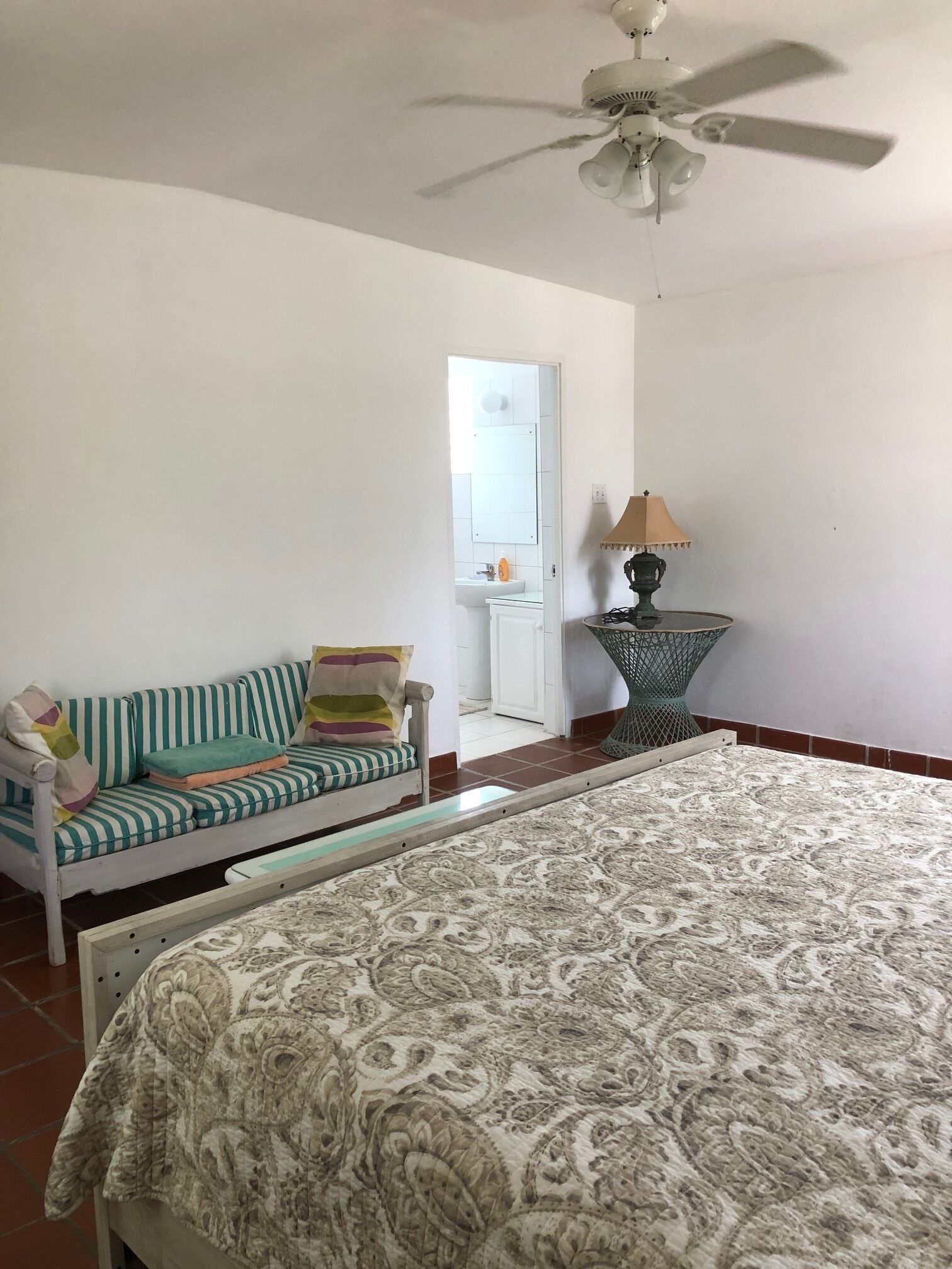 A bedroom with a bed, a sofa, a ceiling fan, a lamp, and a bathroom visible through an open door.