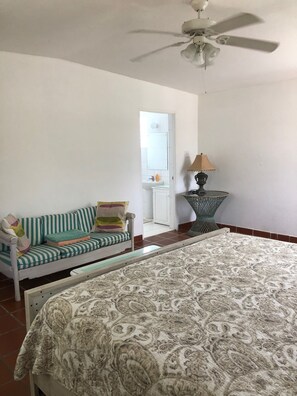 A bedroom with a bed, a sofa, a ceiling fan, a lamp, and a bathroom visible through an open door.