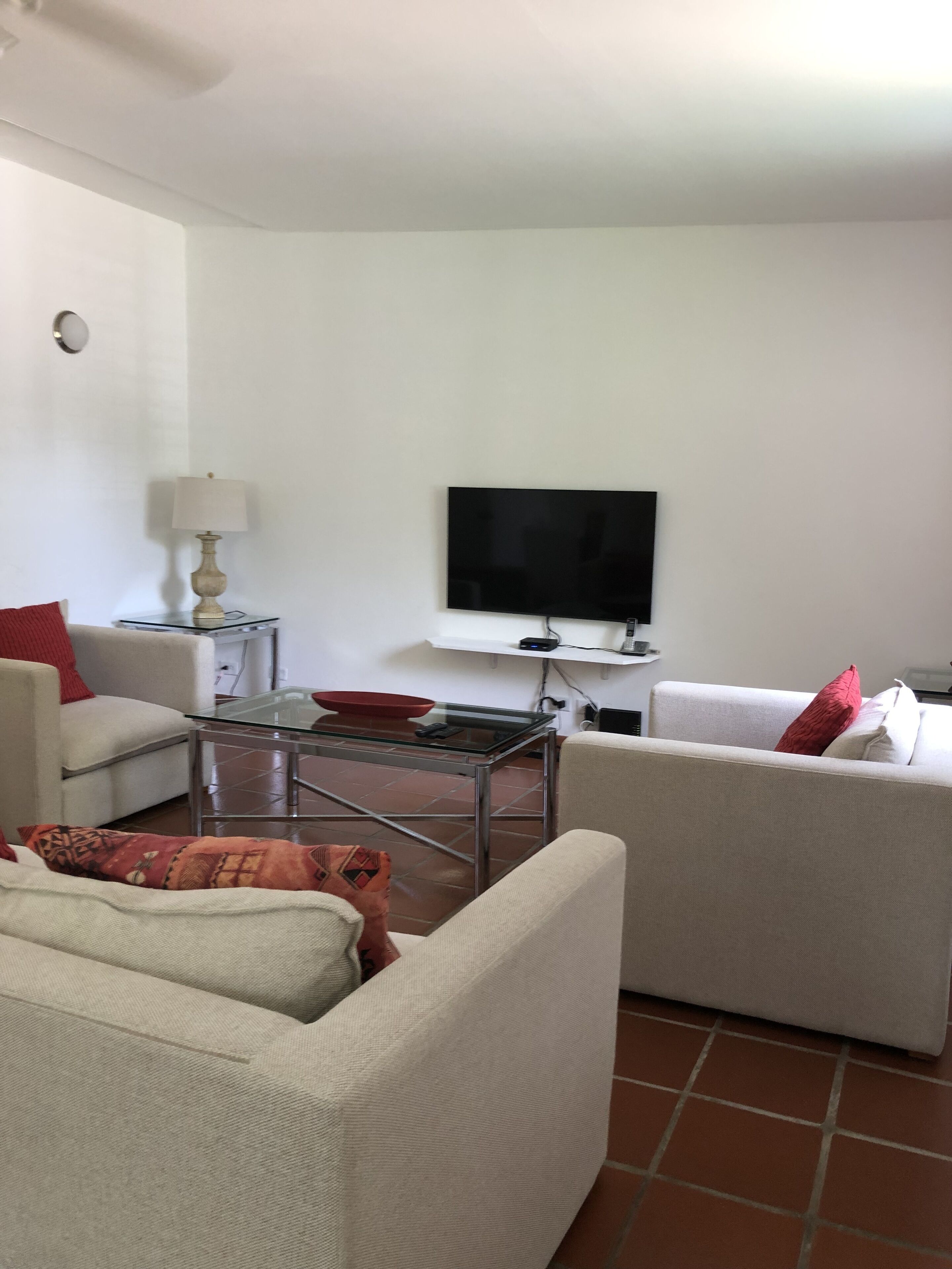 Un salon avec deux canapés blancs, une table basse en verre, un téléviseur fixé au mur et une lampe sur une table d’appoint.