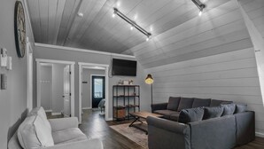 Una sala de estar moderna con un sofá gris, un sillón blanco y un sofá gris oscuro. El piso es de madera, hay una estantería y un televisor de pantalla plana montado en la pared.