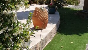Un jardín con diversas plantas, una maceta grande y un edificio con contraventanas azules.