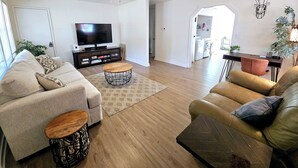 A modern living room with two sofas, a coffee table, and a TV. There is a ceiling fan, a kitchen in the background, and a dining area visible through an open door. - Madrona's Guesthouse- Spacious 2BR with Huge Fenced Yard (North Charleston)