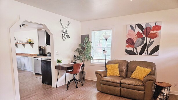 A modern living room with a brown leather sofa, a dining table with two chairs, a potted plant, and a kitchen in the background. - Madrona's Guesthouse- Spacious 2BR with Huge Fenced Yard (North Charleston)