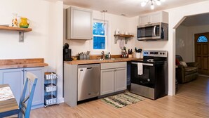 A compact kitchen with a dishwasher, stove, microwave, and a small dining area with a chair and table. - Madrona's Guesthouse- Spacious 2BR with Huge Fenced Yard (North Charleston)