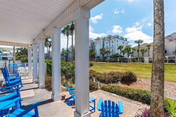 A covered outdoor seating area with blue chairs, white columns, and a view of a building complex with palm trees.