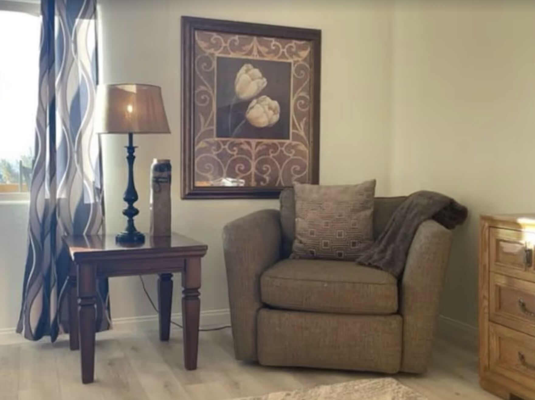 A living room with a brown armchair, a small table with a lamp, a framed picture on the wall, and a wooden dresser.