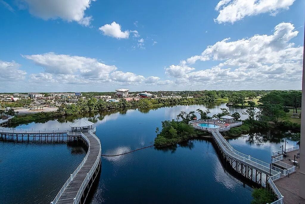 A waterfront area with a pier, a small island with a pool, and a cityscape in the background.