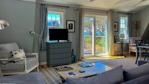 Una sala de estar con televisor sobre un aparador, un sofá, una mesa de centro con revistas y vistas al exterior a través de amplios ventanales.