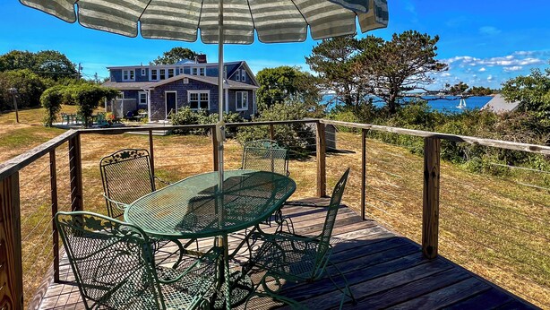 Una terraza de madera con una mesa y sillas de metal, una sombrilla y una casa con vistas al mar al fondo.