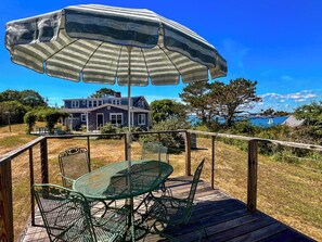 Une terrasse en bois avec une table et des chaises en métal, un parasol, et une maison dont on aperçoit la mer au loin.