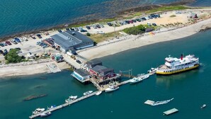 Vista aérea de un puerto deportivo costero con un gran ferry atracado, varias embarcaciones más pequeñas y un aparcamiento con coches.