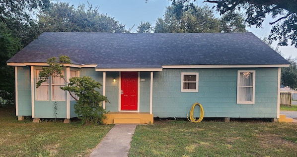 A single-story house with a red door, a small porch, and a yellow hose on the side.