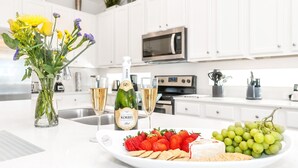 A modern kitchen with white cabinets, a stainless steel sink, and a microwave. There is a glass vase with flowers, a bottle of champagne, and a plate of strawberries and grapes. - Golf Hideaway With Lake View! Modern Decor and Game Room With Multiple Options! #5pr2197 (Davenport)