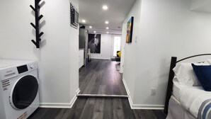 A laundry room with a front-loading washer and dryer, a hallway leading to a bedroom with a bed and blue pillows, and a living area with a couch and a painting on the wall.