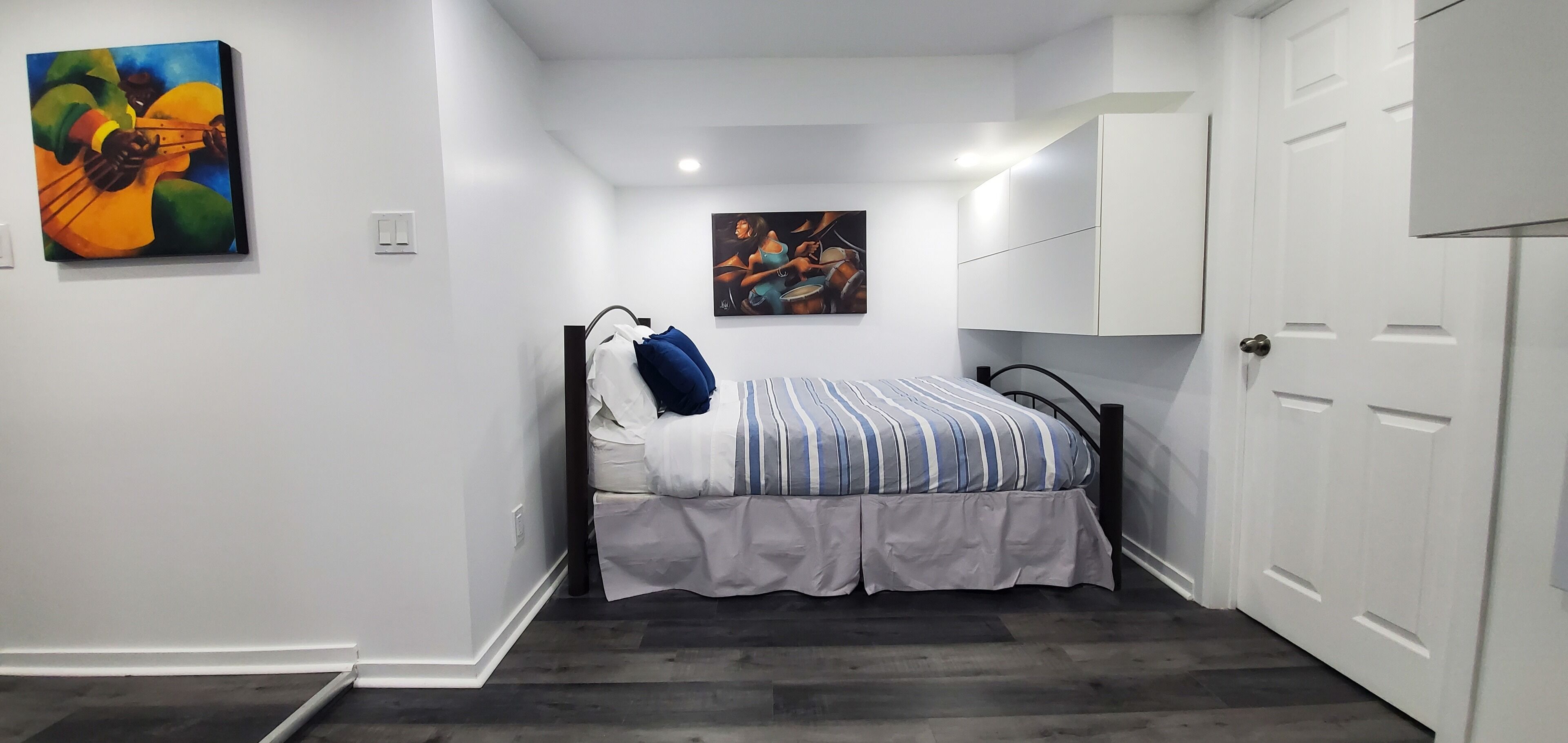 A bedroom with a bed, a wall-mounted guitar painting, and a painting of a woman.