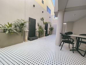 Un estrecho pasillo exterior con suelo estampado, plantas en macetas y una zona de estar con mesa y sillas.