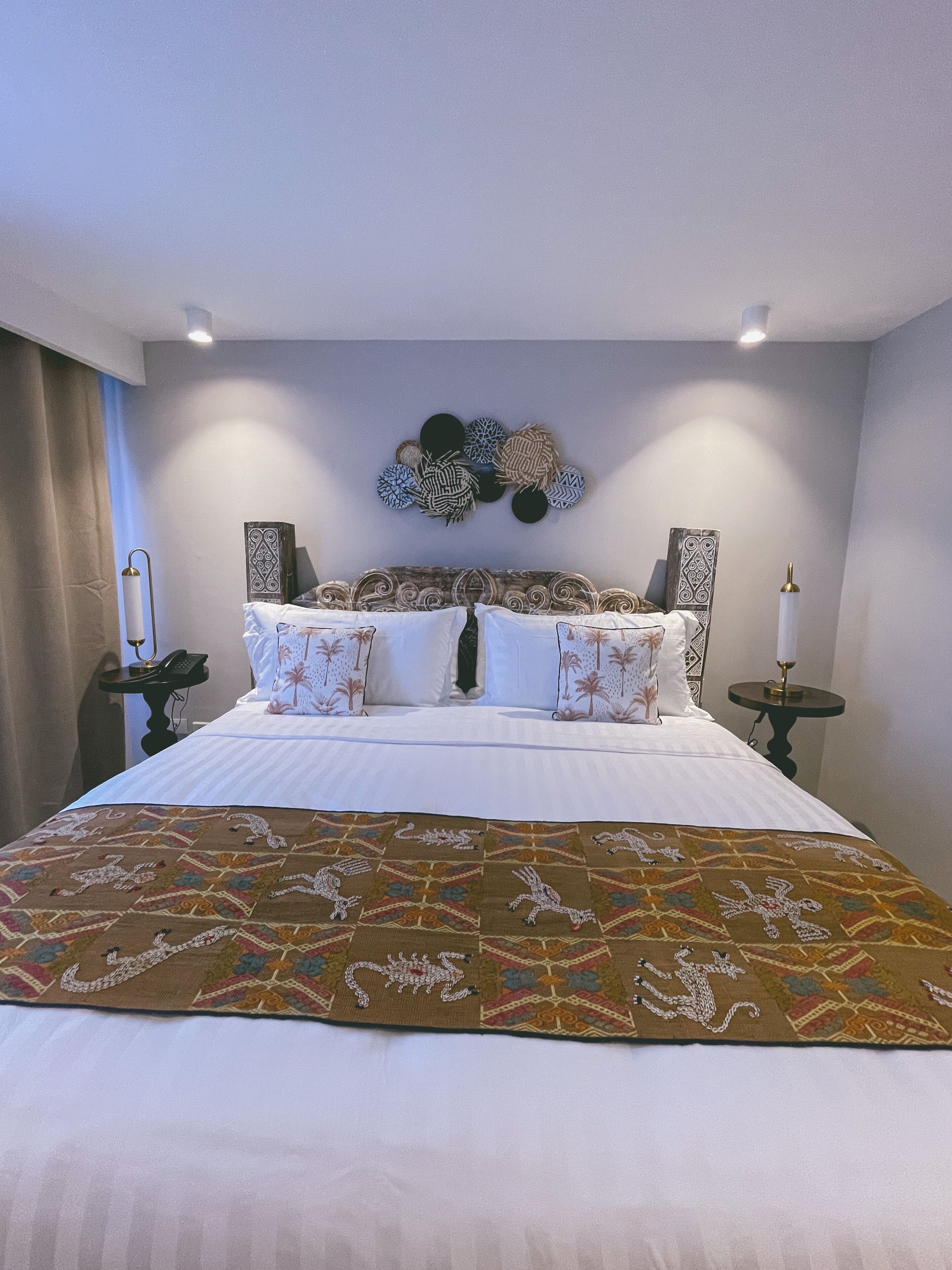 Un lit bien fait, recouvert d’une couvre-lit à motifs, une œuvre d’art décorative accrochée au mur au-dessus, et deux tables de chevet avec des lampes.