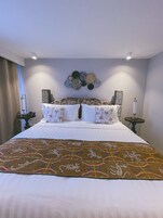 Una cama bien hecha con una colcha estampada, una obra de arte decorativa sobre ella y dos mesitas de noche con lámparas.