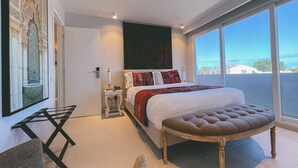 A modern hotel room with a large bed, a sitting area with a chair, a small table, and a large window offering a view of the city.