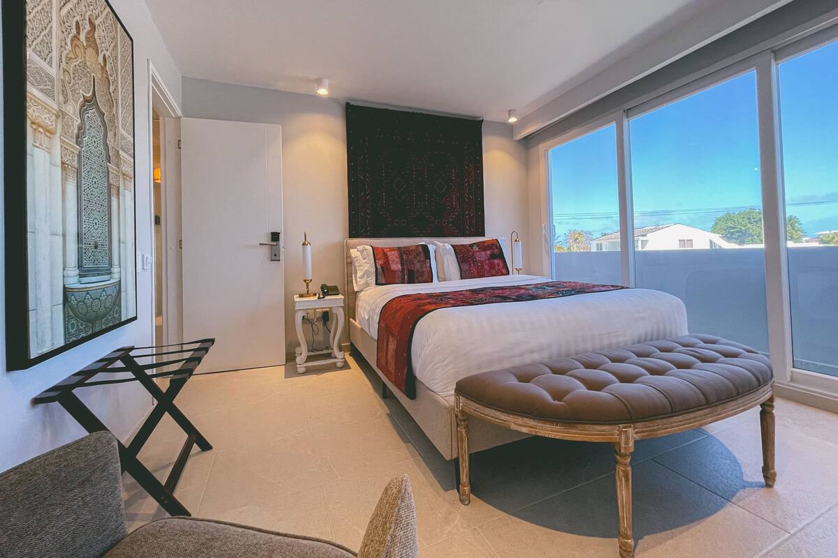 Una habitación de hotel moderna con una cama grande, una zona de estar con una silla, una mesita y un amplio ventanal con vistas a la ciudad.