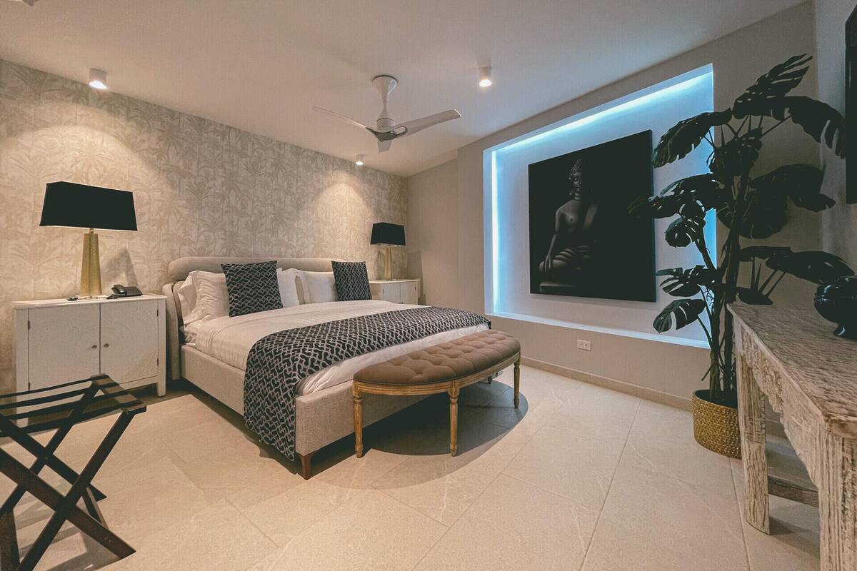 Un dormitorio moderno con cama, mesitas de noche, un banco, ventilador de techo y una ventana grande con una planta.