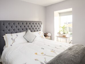 A bedroom with a tufted headboard, a bed with floral bedding, a bedside table with decorative items, and a window with a view of greenery.