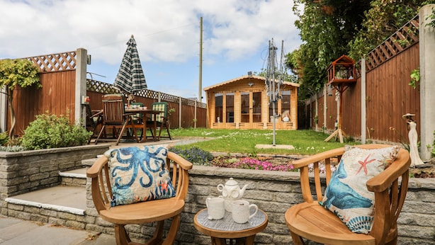 Una terraza de madera con dos sillas y una mesita, una cabaña de madera al fondo y una valla de madera que delimita el área.