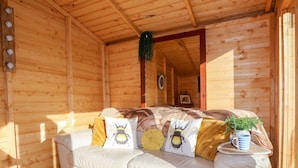 Interior acogedor de una cabaña de madera con un sofá de cuero blanco decorado con almohadones con motivos de abejas, una planta pequeña en una mesita auxiliar y un techo de madera.