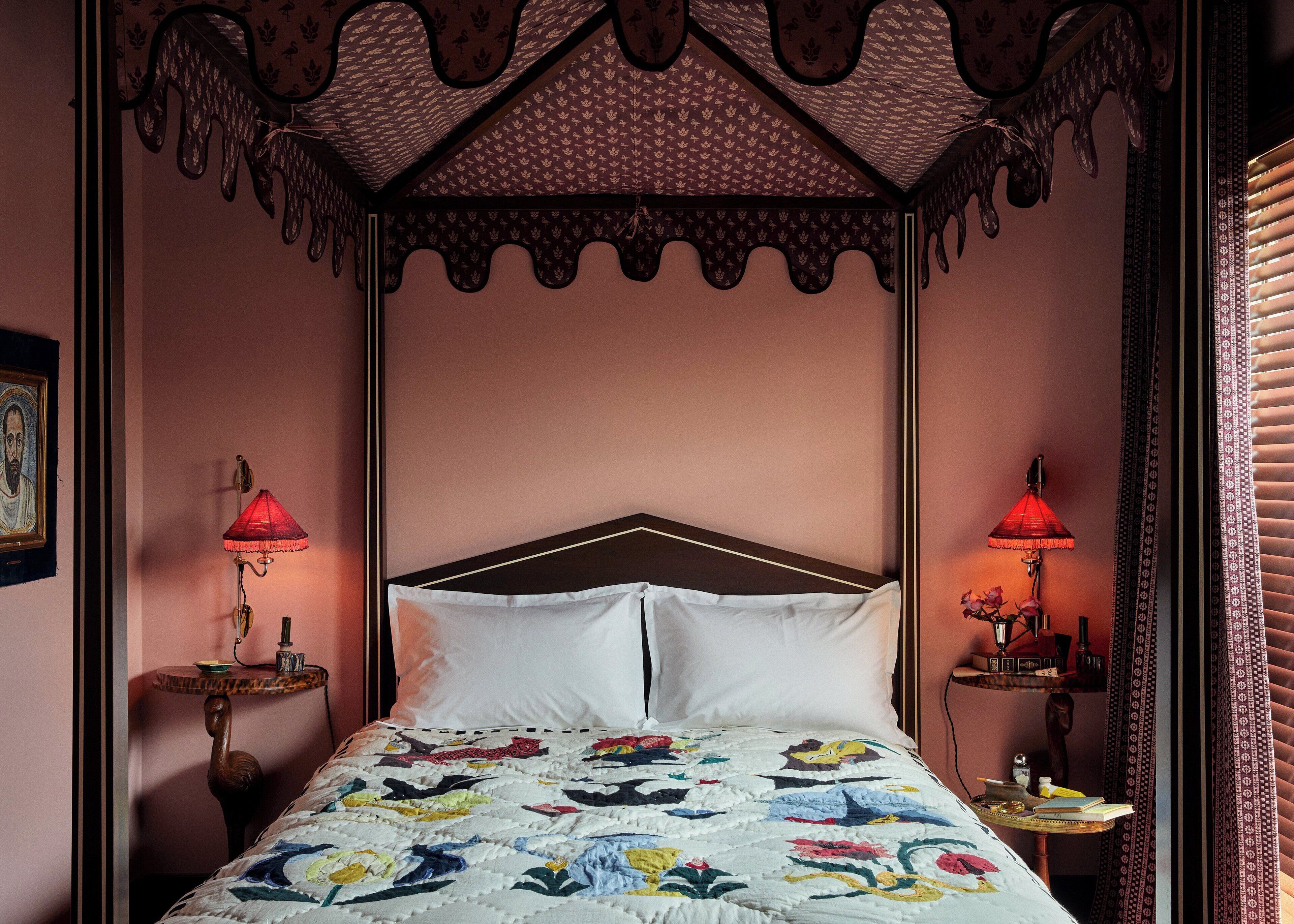 A bedroom with a canopy bed, two bedside tables, a framed picture on the left wall, and a red lamp on each side of the bed.